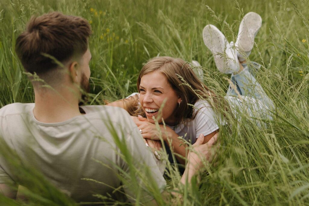 Woman lays on her stomach in the grass with her head on her fiance's lap.