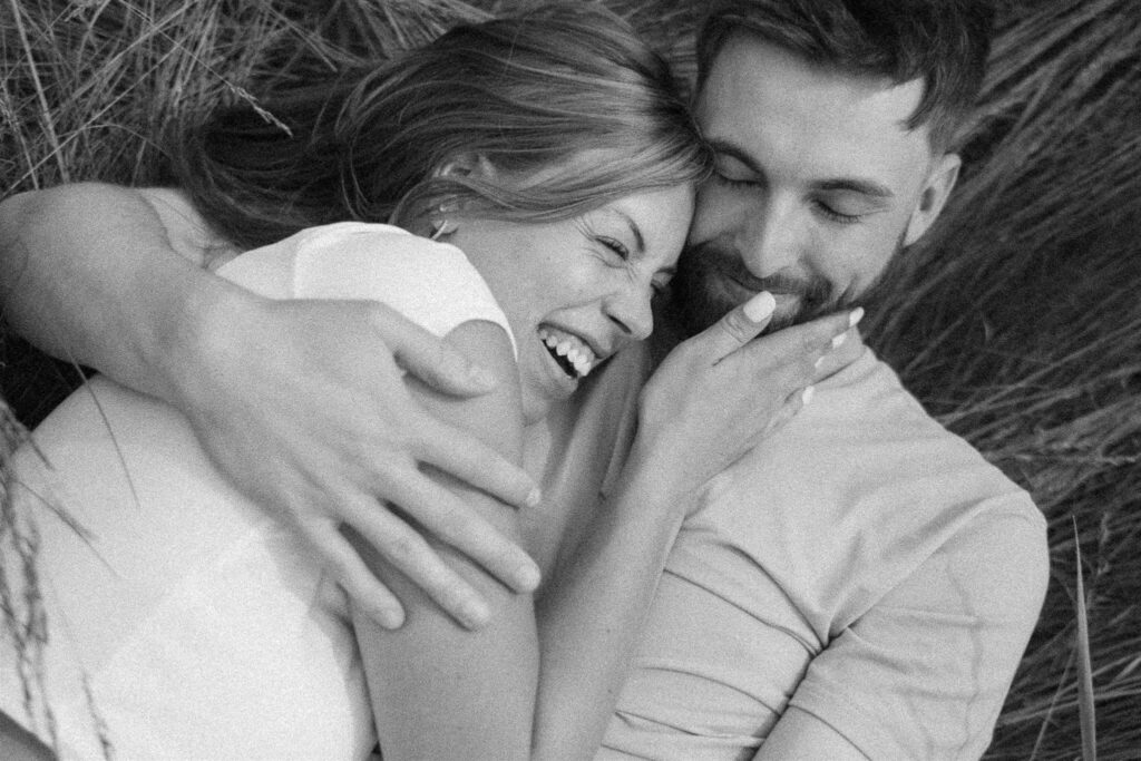 Woman wearing classic white tee and light wash denim jeans laughs and cups her fiance's face as he holds her close