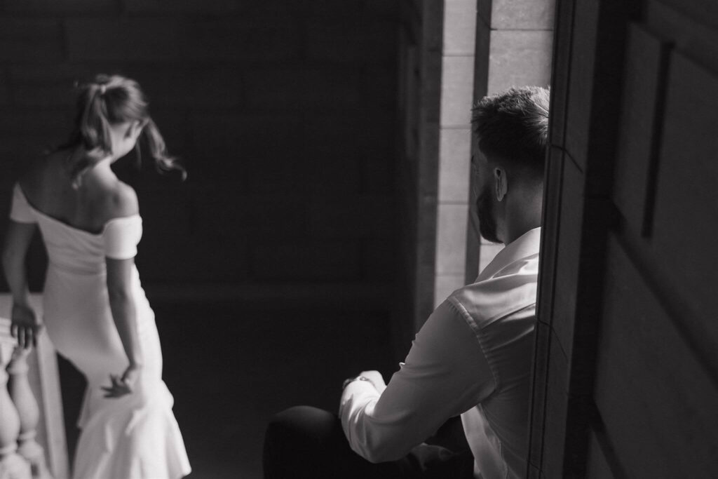 Man watches his fiancee descend down the stairwell in her white dress in engagement photos