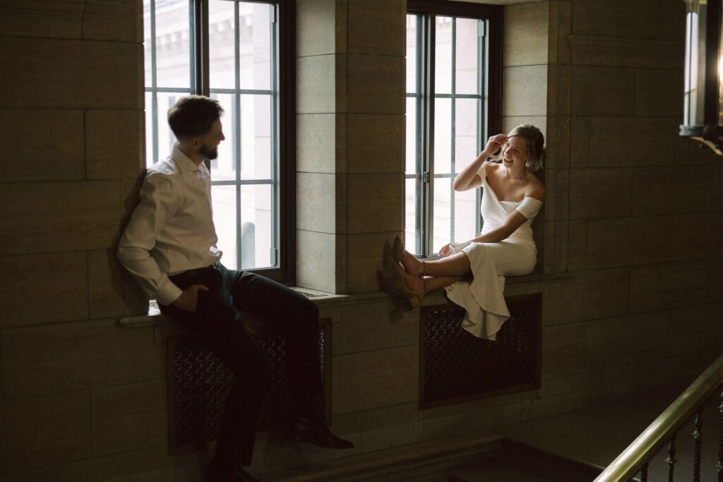 Engaged couple looks at watch other as they sit on the windowsill of classic venue in their engagement photos