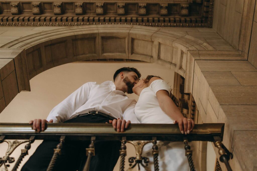 Man and woman kiss as they both grip the banister of elegant engagement photos venue