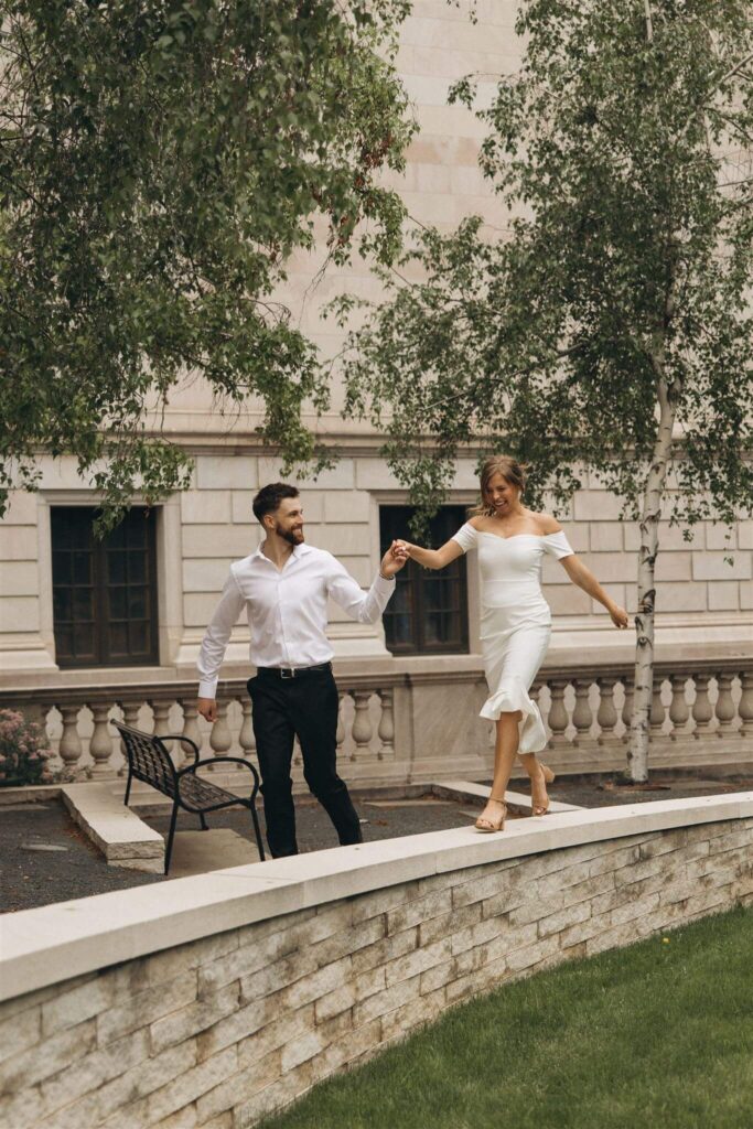 Man in white dress shirt and black dress pants holds his fiance's hand as she walks along a ledge in heels