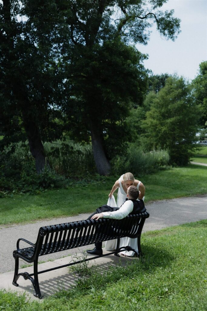 Bride leans down to kiss groom sitting on park bench