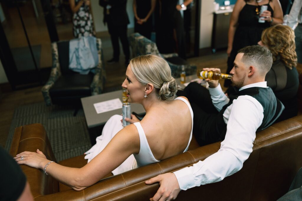 Bride and groom enjoy drinks on leather couch during cocktail hour