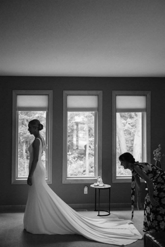 Mother of the bride fixes bride's train as she stands in front of living room windows