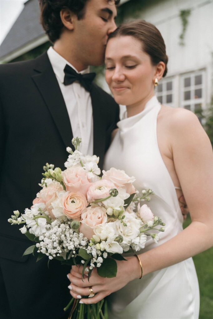 Groom kisses the top of bride's head as she looks down at her bridal bouquet