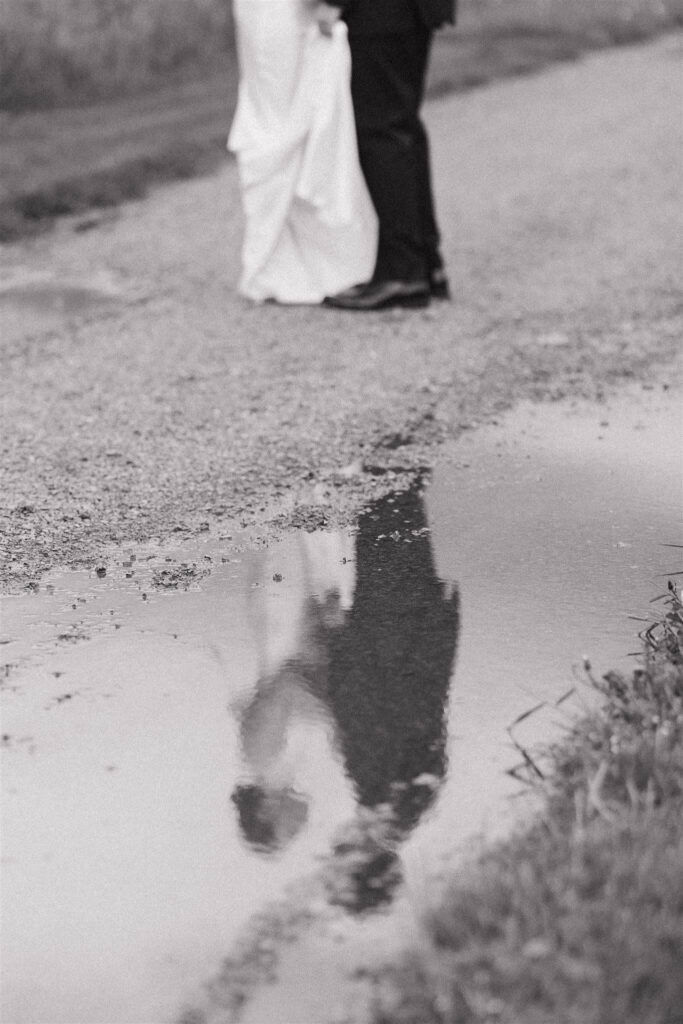 Bride and groom are reflected in a puddle on the gravel