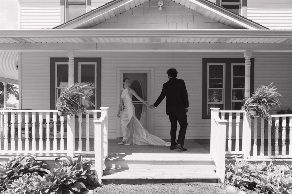 Bride and groom hold hands on front porch of farmhouse wedding venue