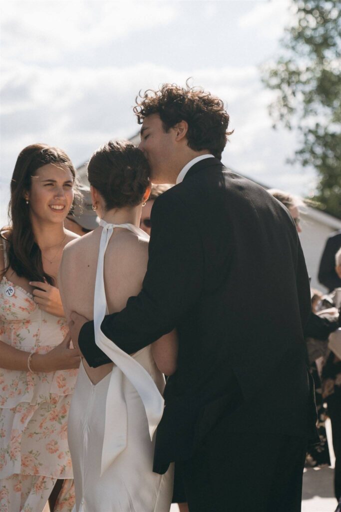 Groom lays a kiss on bride's head as they socialize with wedding guests during cocktail hour