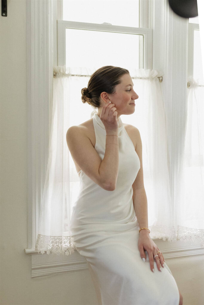 Bride sits on windowsill while she fixes her earrings