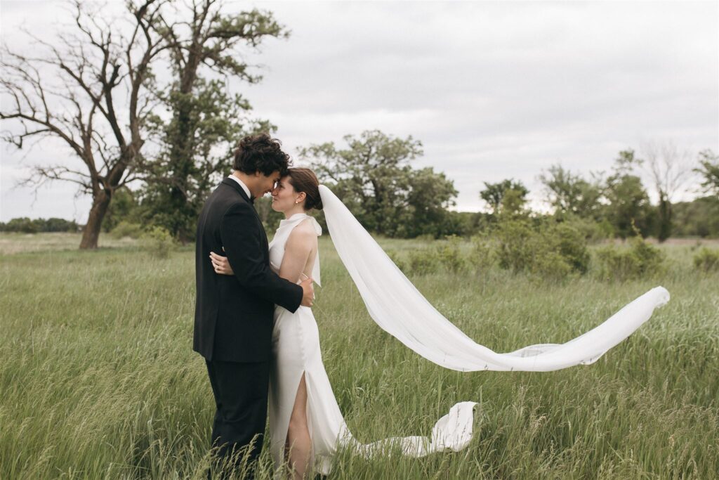 Bride and groom smile at each other in Minnesota meadow as the wind whips her cathedral veil