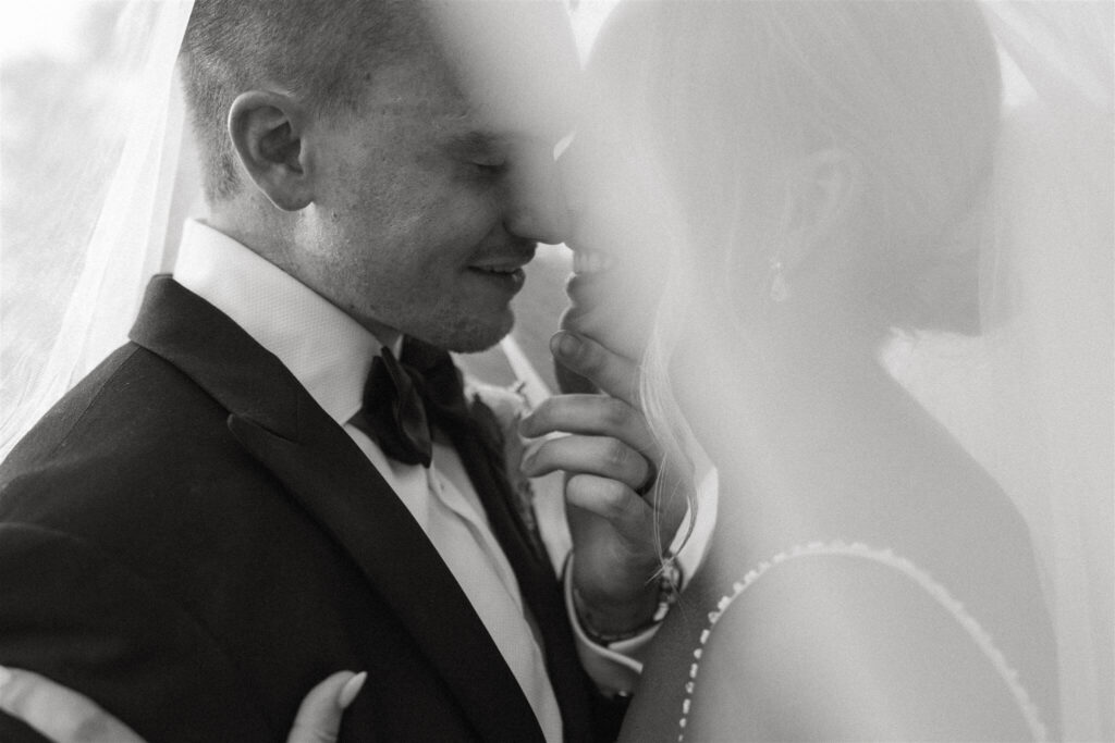 Groom cups bride's chin as he goes in for a kiss under the shelter of her veil