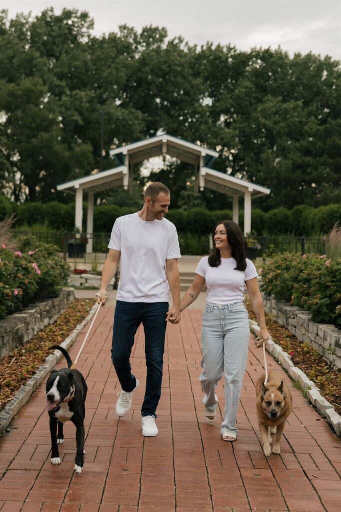 Engaged couple walk hand in hand with their two dogs on a leash in park engagement photo shoot