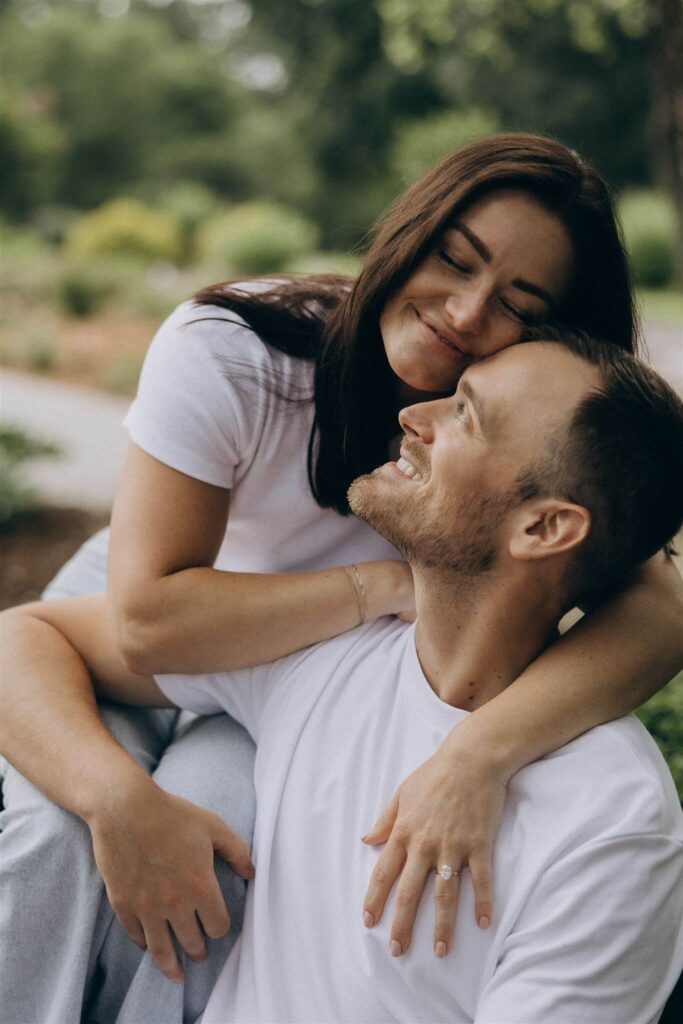 Man smiles up at his fiance as she puts a hug around him from behind