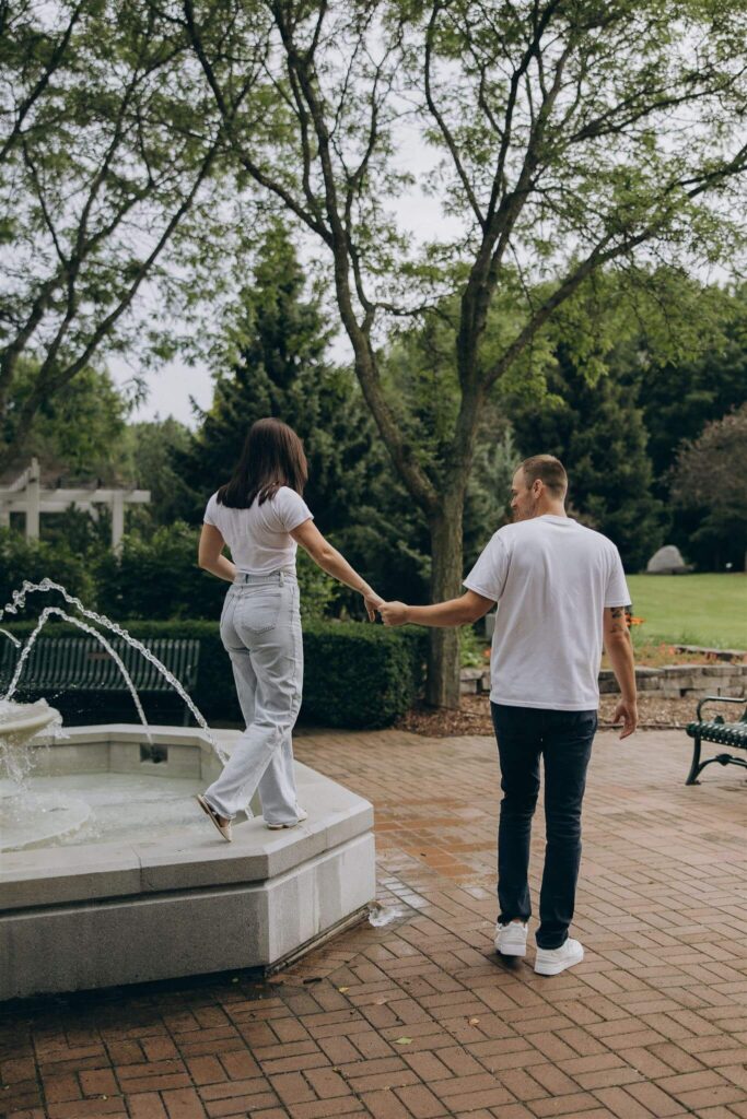 Man holds woman's hand as she balances on the ledge of a park fountain