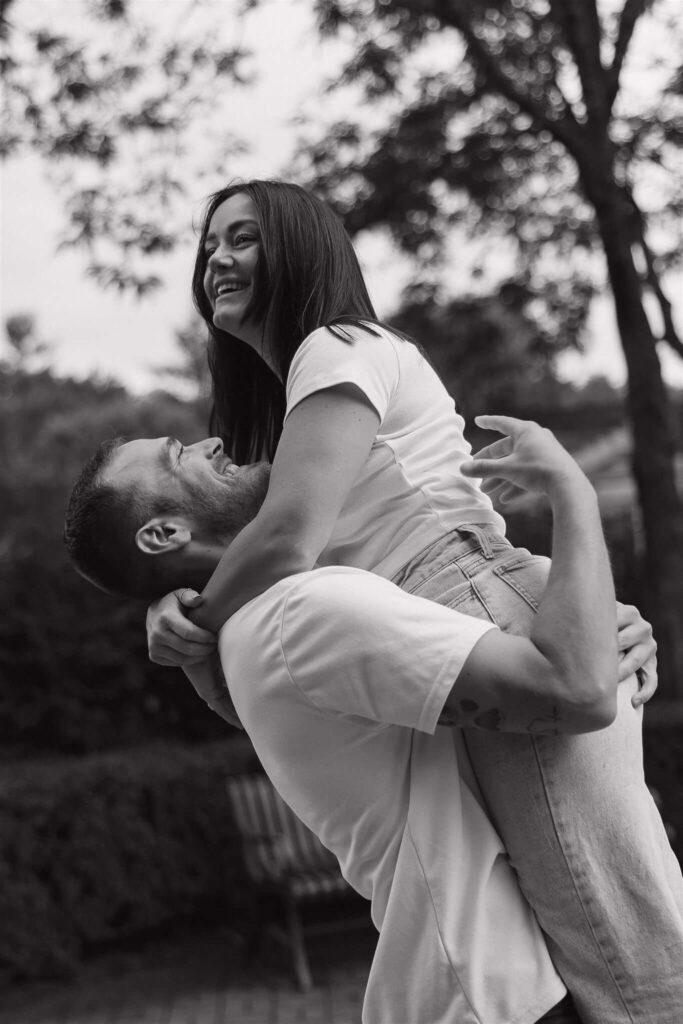 Man wearing white tee and denim jeans lifts his fiance up into the air in Twin Cities engagement photo shoot