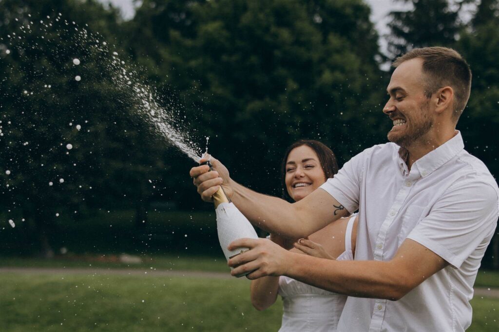 Man pops the cork off of champagne bottle while his fiancee claps