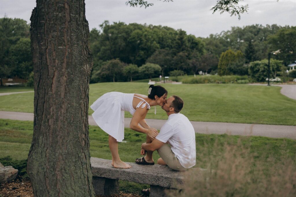 Woman stands on park bench and leans down to kiss her fiance in their engagement photo shoot