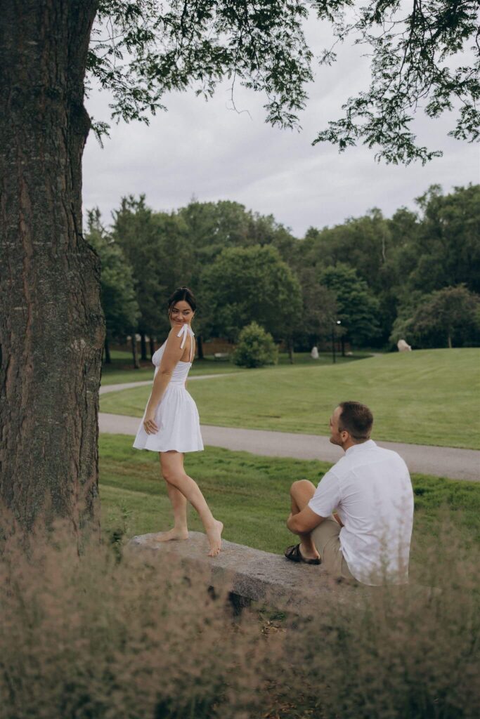 Man looks on as his fiance dances barefoot on a park bench