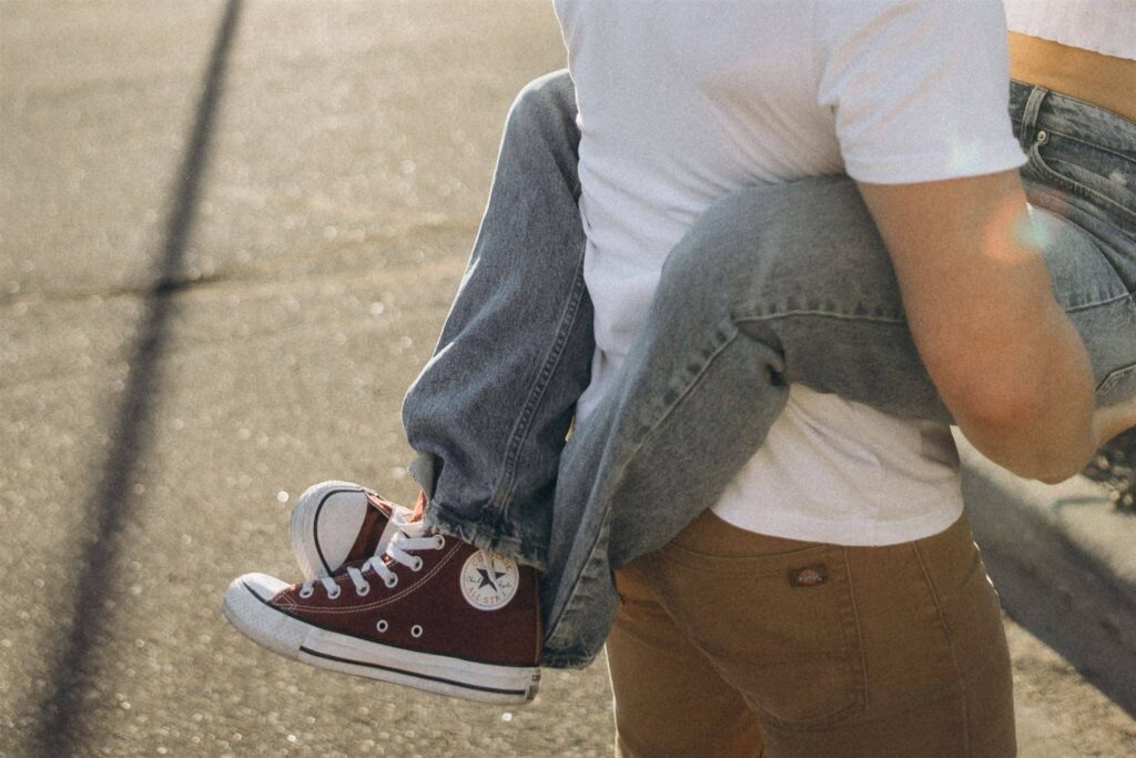 Woman wearing red Converse and denim blue jeans wraps her legs around her fiance's waist.