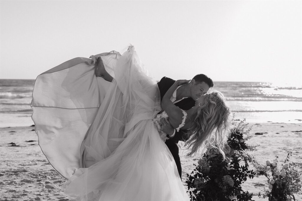 Groom grins at bride as he dips her in a romantic beach kiss