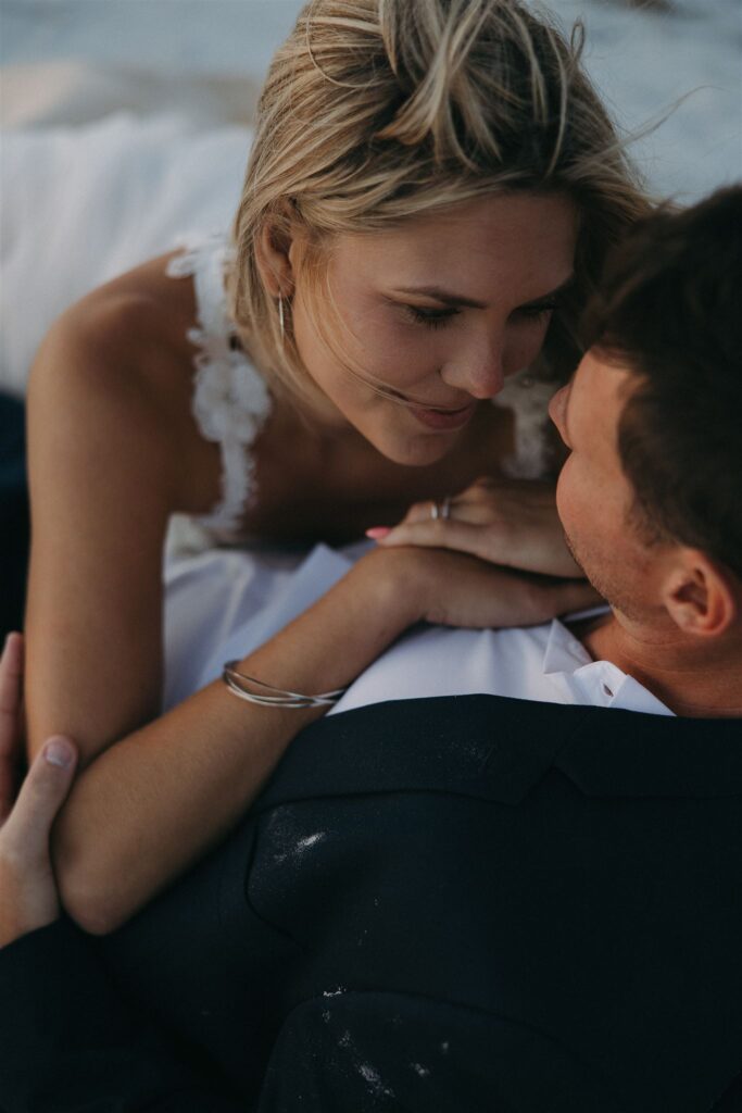 Bride smiles at groom as they lay in the sand, chest to chest