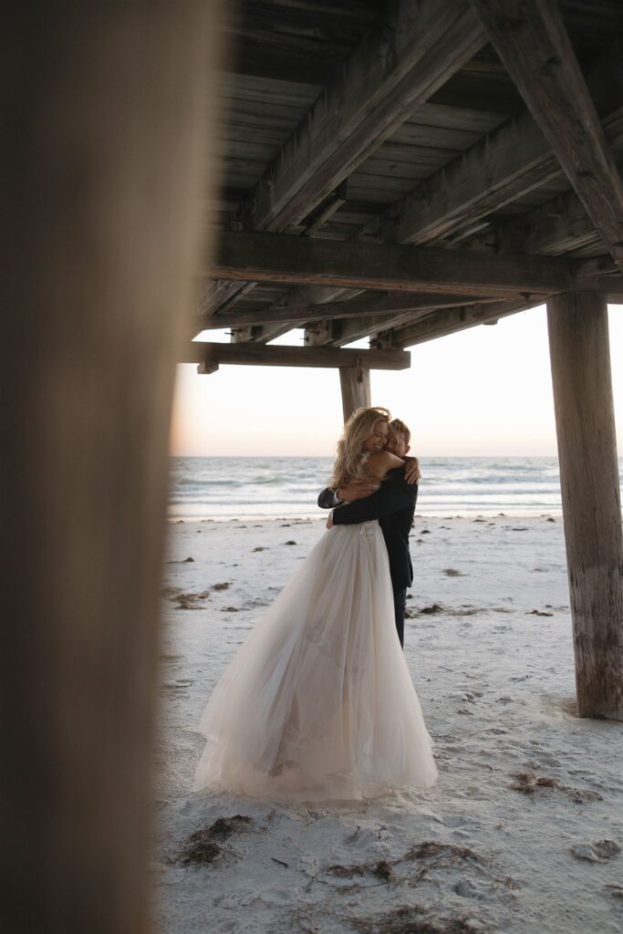 Bride and groom hug under Florida pier at beach destination elopement