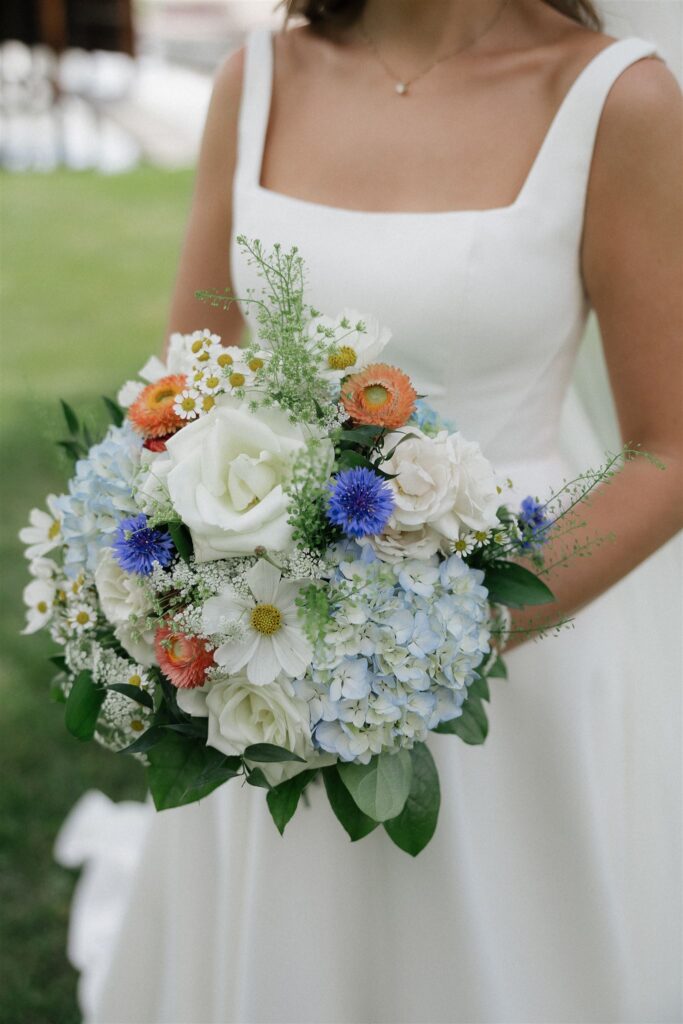 Bride holds a bouquet of white roses and daisies, blue hydrangeas, and assorted other flowers