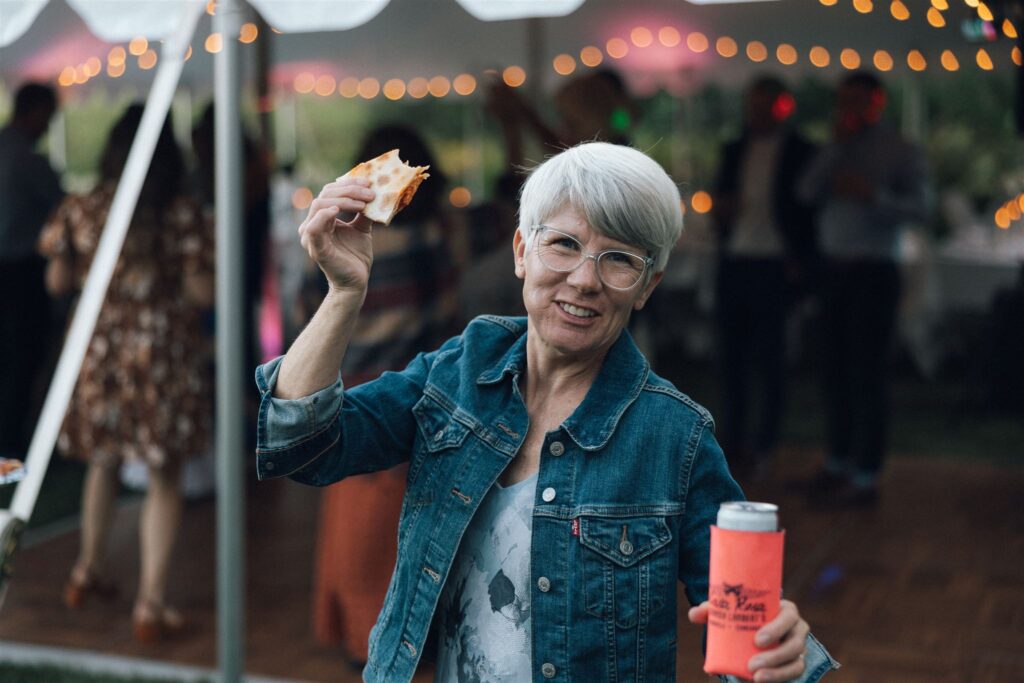 Mother of the bride holds a beer and her pizza up in the air