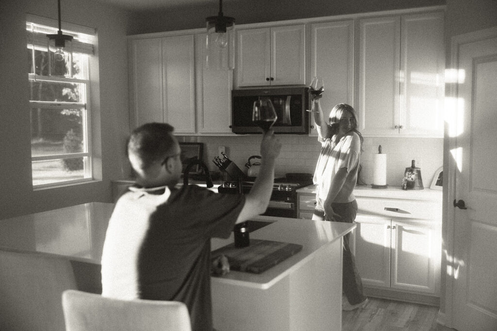 Woman dancing around kitchen raises her wine glass in the air while husband looks on from the kitchen island