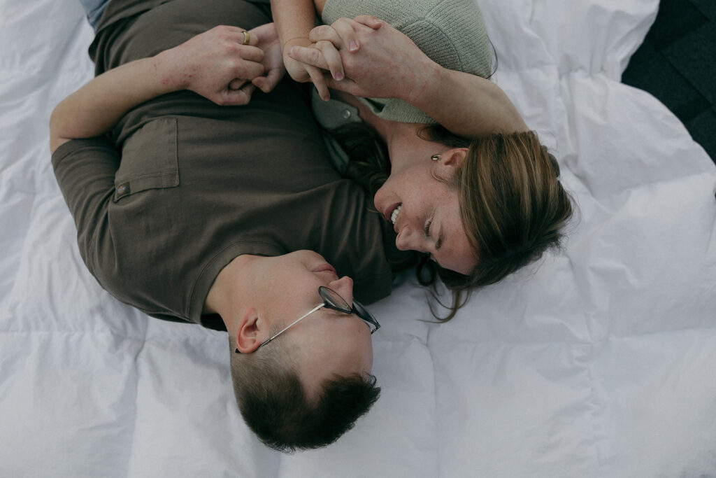 Married couple lays on white duvet with fingers intertwined