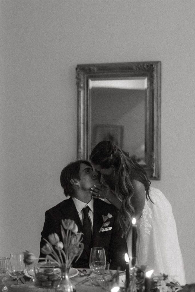 Bride stands behind groom and leans into kiss him where he sits