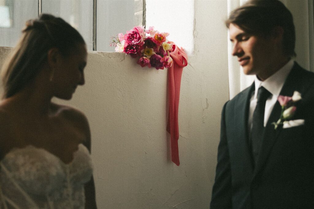 Bride and groom share intimate moment in the foreground while bridal bouquet rests on the windowsill