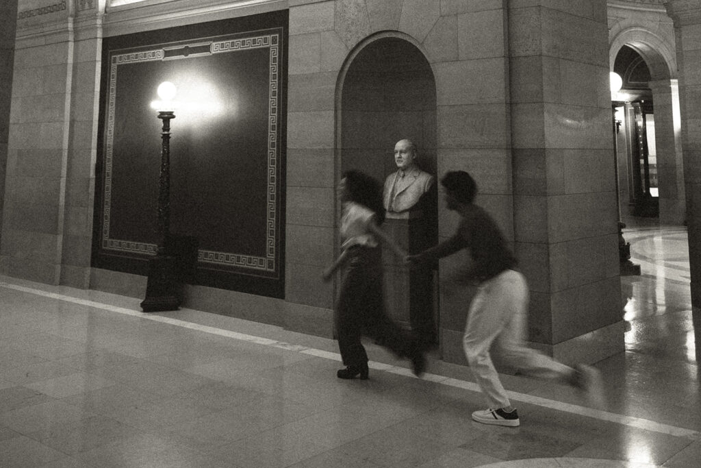 Shubhi and Abhi holding hands beneath grand architecture, cinematic engagement feel