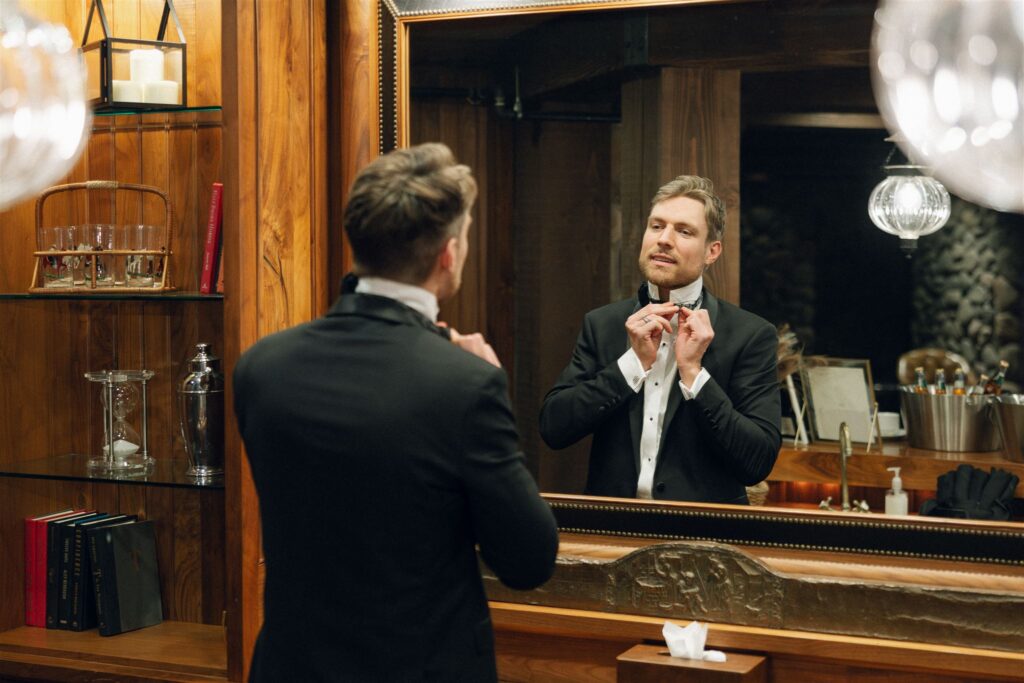 Groom adjusting his tie in the mirror inside the warm wood-accented groom’s quarters at Edward Anne Estate