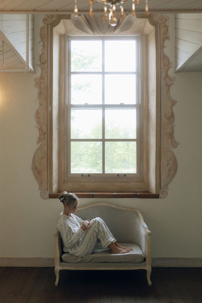 Bride in pajamas gazing out the window from chaise lounge in bridal suite.