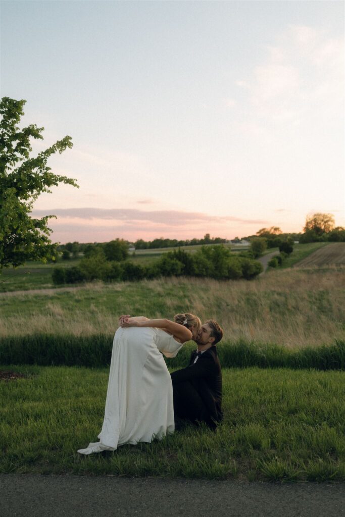 Bride stoops down to kiss groom where he sits in the grass, framed by trees and sky.