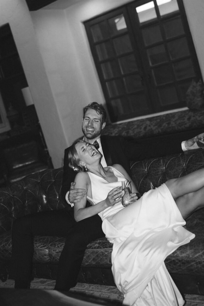 Bride and groom laugh while relaxing on a vintage leather couch in the venue lounge.