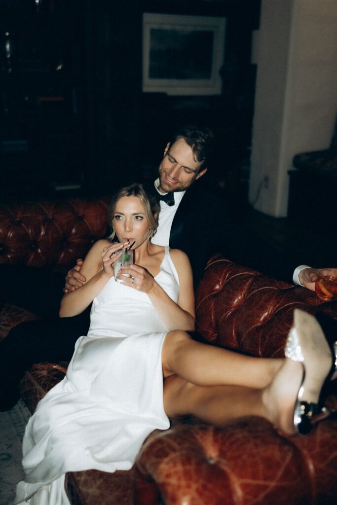 Bride lounges in groom’s arms on a leather couch, sipping a drink in her reception dress.