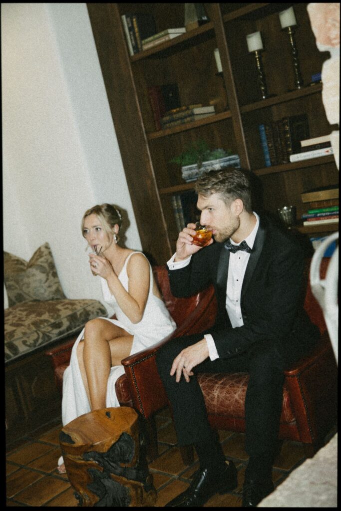 Bride and groom sip cocktails in the venue library, surrounded by bookshelves.