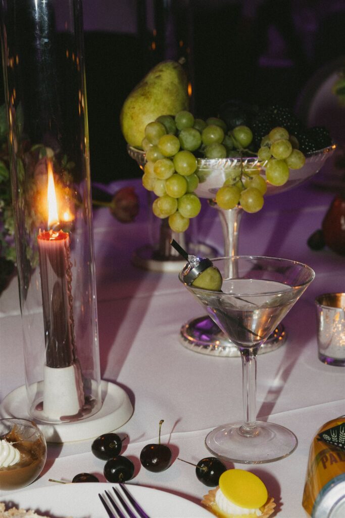 Reception flat lay of candlelit table with grapes, figs, and drinks in elegant glassware.