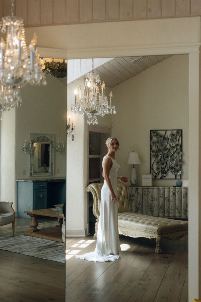 Bride standing in natural light beside a full-length mirror under sparkling chandeliers.
