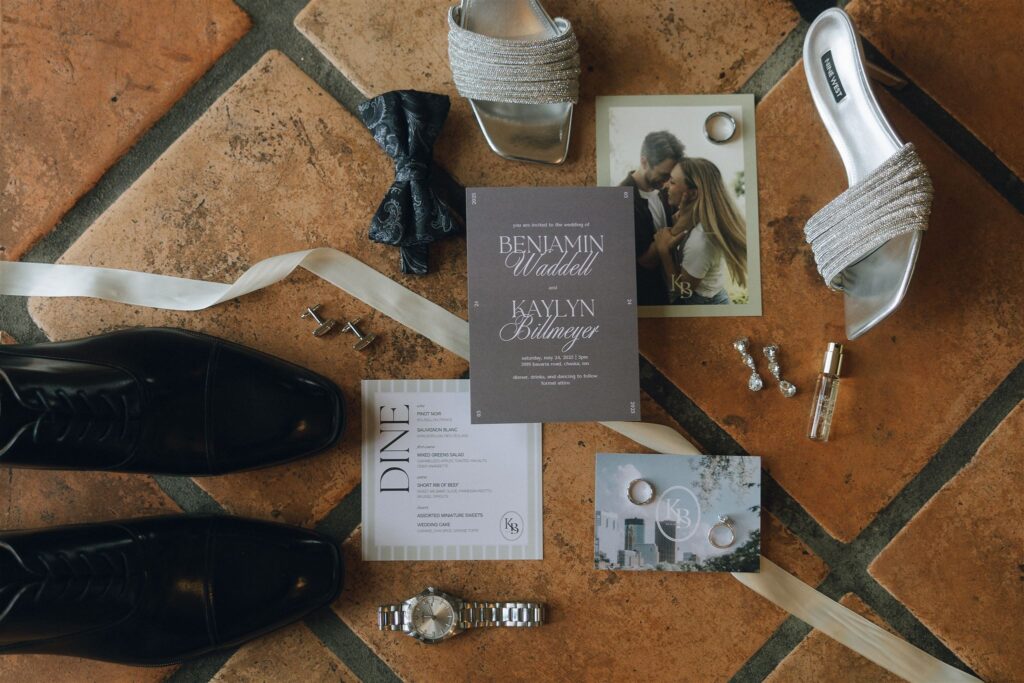Flatlay of wedding details including invitations, rings, and accessories on tiled floor