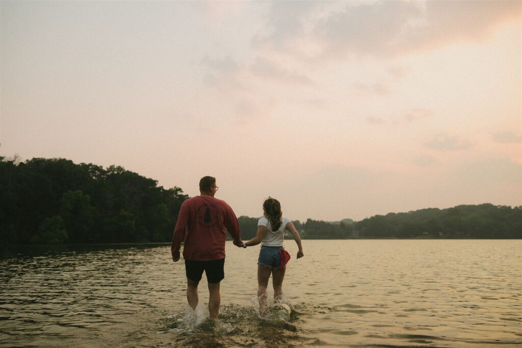 Couple walking hand-in-hand into the shallow lake water, splashing as they go for summer engagement photos