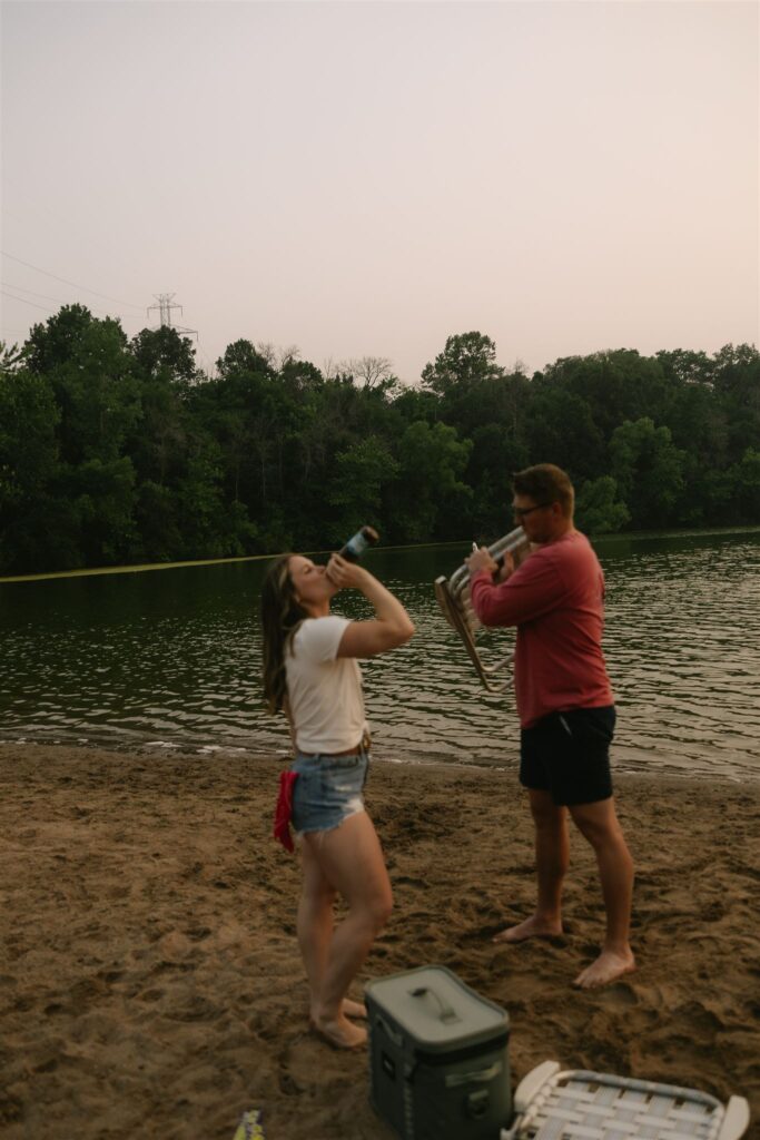 Woman drinking from a beer bottle while man laughs nearby on the beach for summer engagement photos