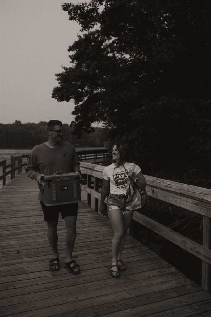 Couple walking down the boardwalk, man carrying a cooler, woman holding a folding chair for summer engagement photos