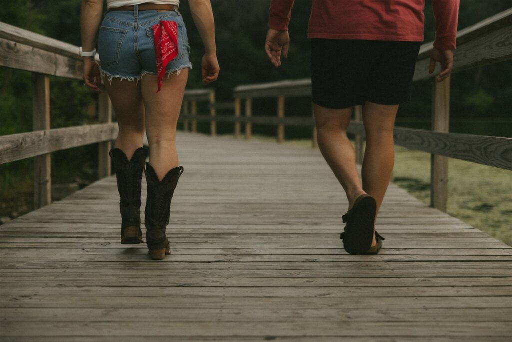 Couple walking side by side down a wooden boardwalk, her in denim shorts and boots, him in red shirt.