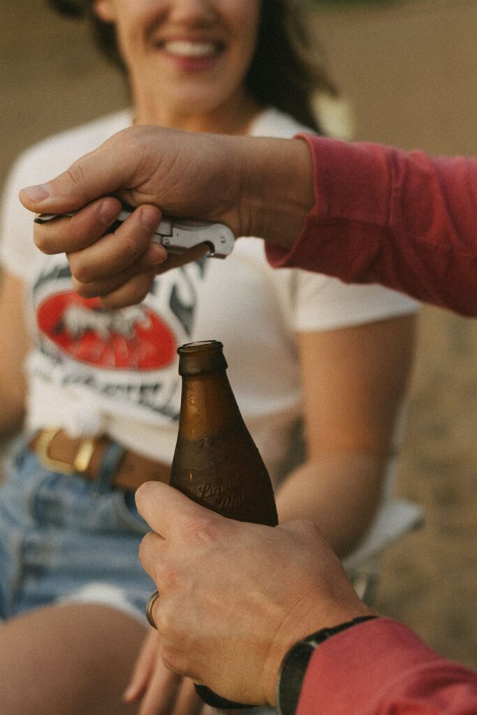 Close-up of woman’s hand reaching for a beer bottle as man opens it.
