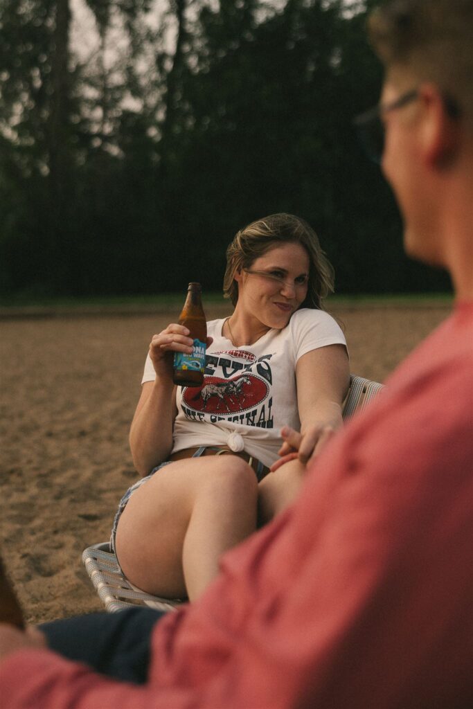 Woman sitting in a beach chair, smiling at man in the foreground while holding a beer.