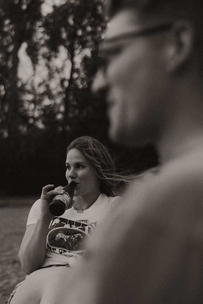 Woman smiling with a beer in hand while man sits beside her out of focus for summer engagement photos
